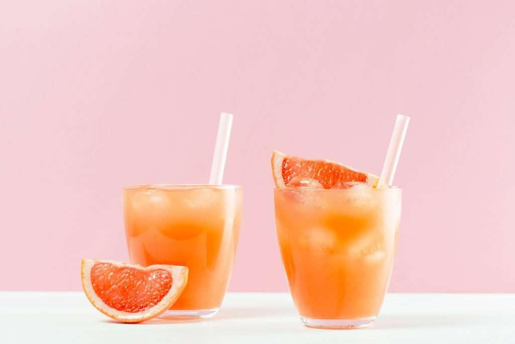Two glasses of grapefruit juice with ice and straws, embodying the principles of Naturopathic Medicine, with a pink backdrop.