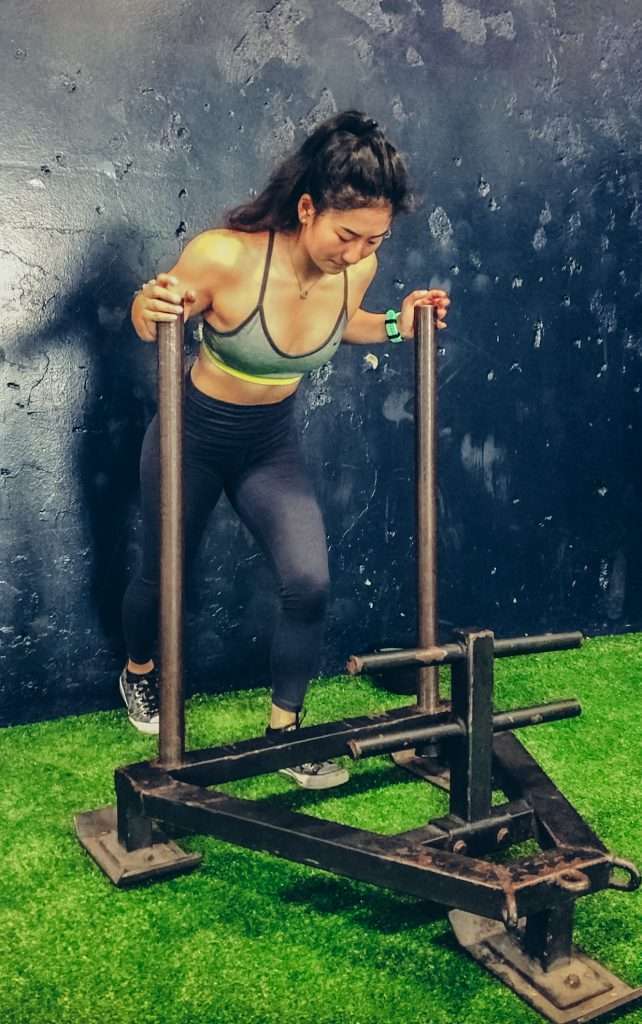 Woman pushing a weighted sled on an indoor turf surface, showcasing the strength developed through functional medicine practices.

