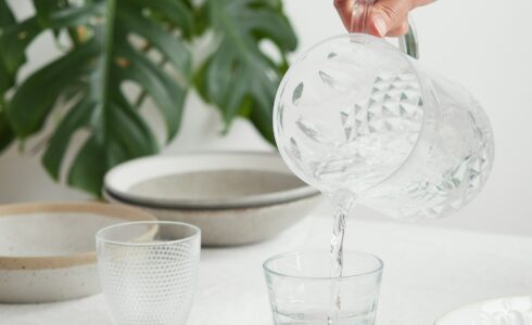 A hand pours water from a clear, patterned pitcher into a glass, emphasizing the importance of hydration. Another glass and several plates are on a table covered with a white cloth, with a large green plant in the background, adding to the natural health ambiance.