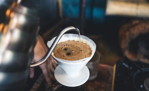 A close-up of a barista in Meridian pouring hot water from a gooseneck kettle into a white ceramic dripper on a stand, making pour-over coffee. The image captures the hand pouring the water and the coffee grounds blooming in the filter, creating a moment that feels warmly connected to natural health.