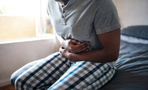 A person sits on a bed, holding their stomach with both hands, clearly displaying discomfort. They are wearing a grey shirt and checkered pants, hinting at the possible struggle with IBS while seeking relief through naturopathic medicine.