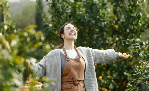 A person wearing a brown apron and a gray sweater stands outdoors in an orchard in Meridian, ID, smiling with arms outstretched thanks to BHRT and functional medicine.