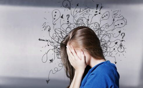 A person with long hair, wearing a blue shirt, covers their face with hands. Behind them are chaotic doodles representing stress or confusion. Amidst the chaos, the calming properties of L-theanine might offer some solace.